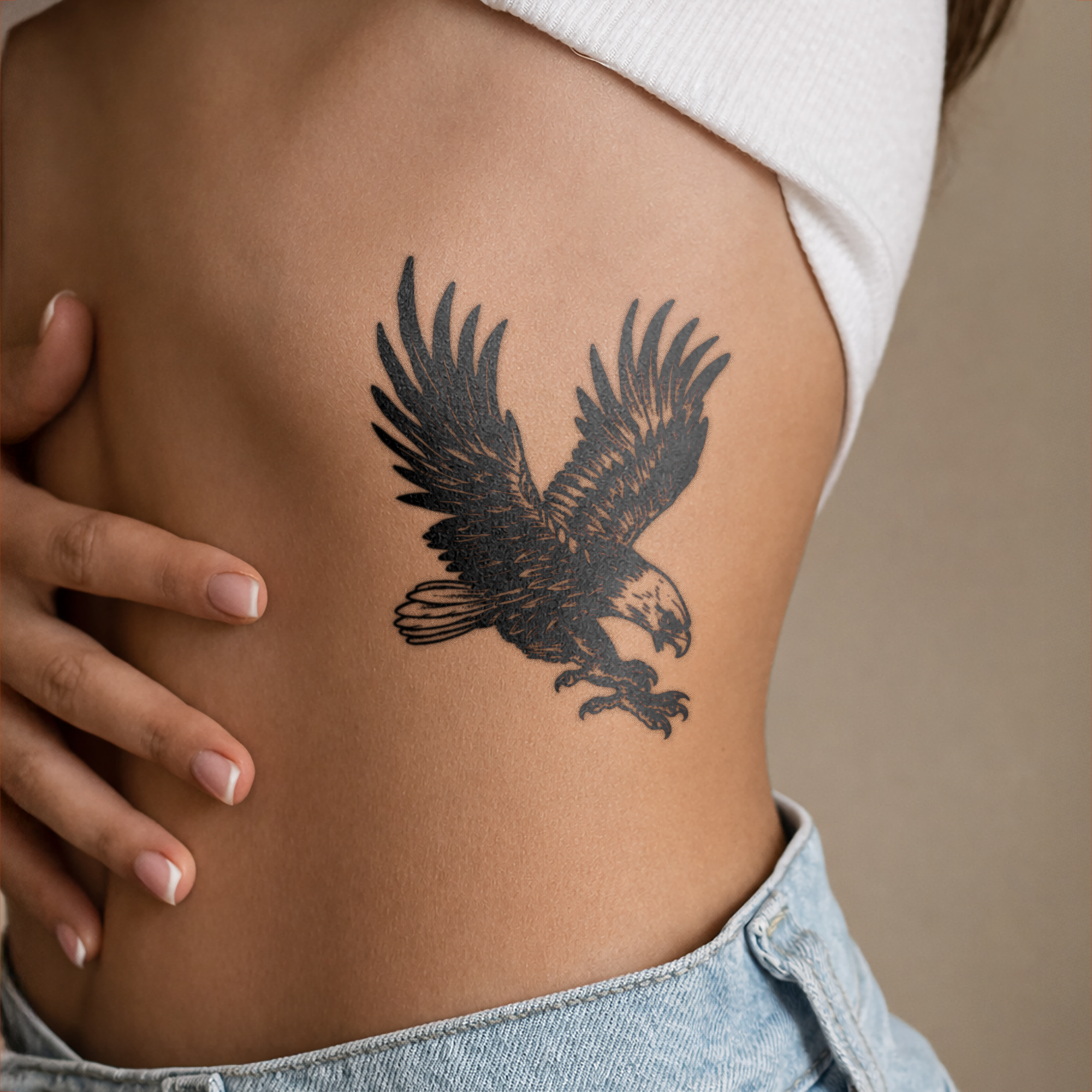 Person with an eagle tattoo on their rib cage, wearing a white top and blue jeans.