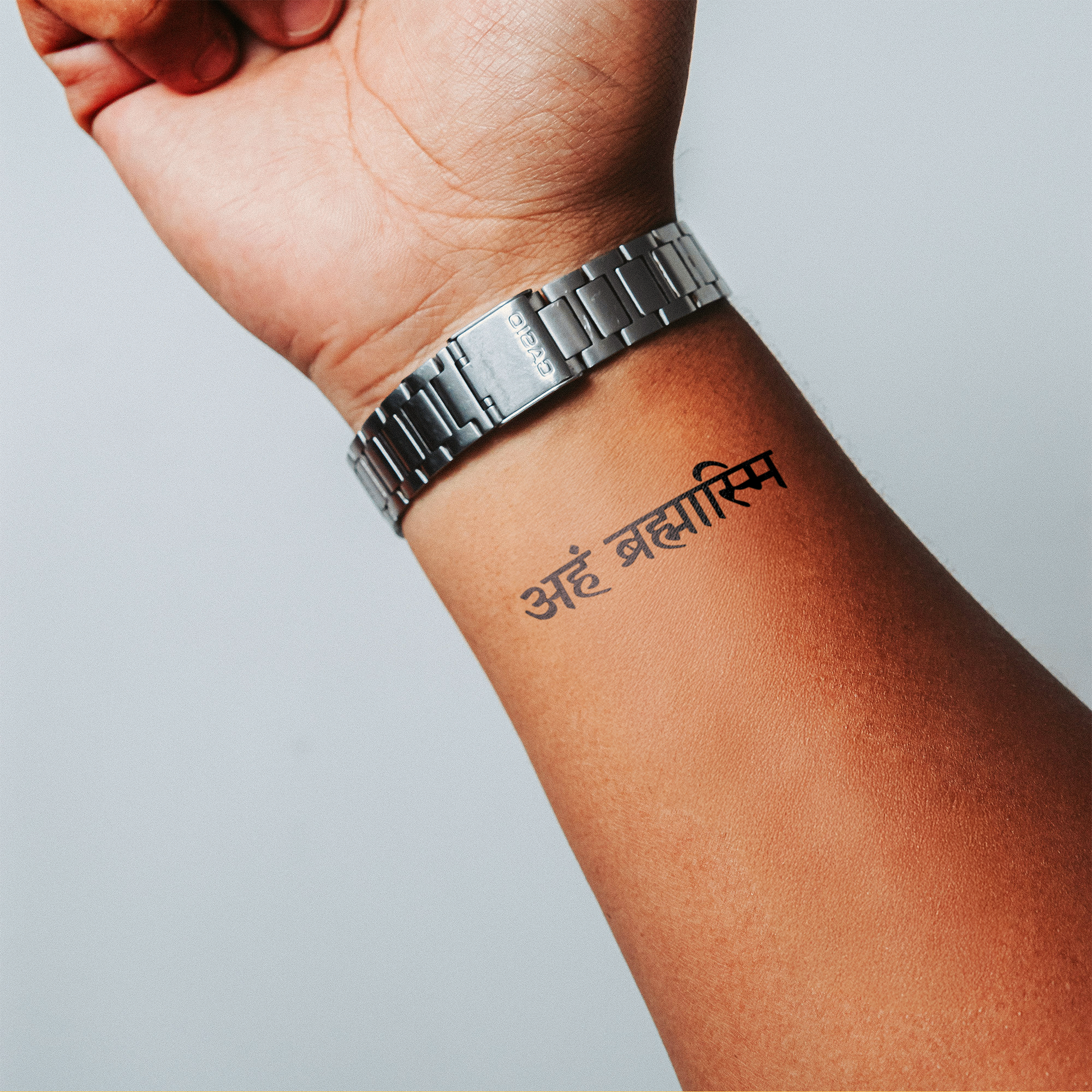 Hand with a silver watch displaying a Hindi phrase tattooed on the wrist against a light gray background