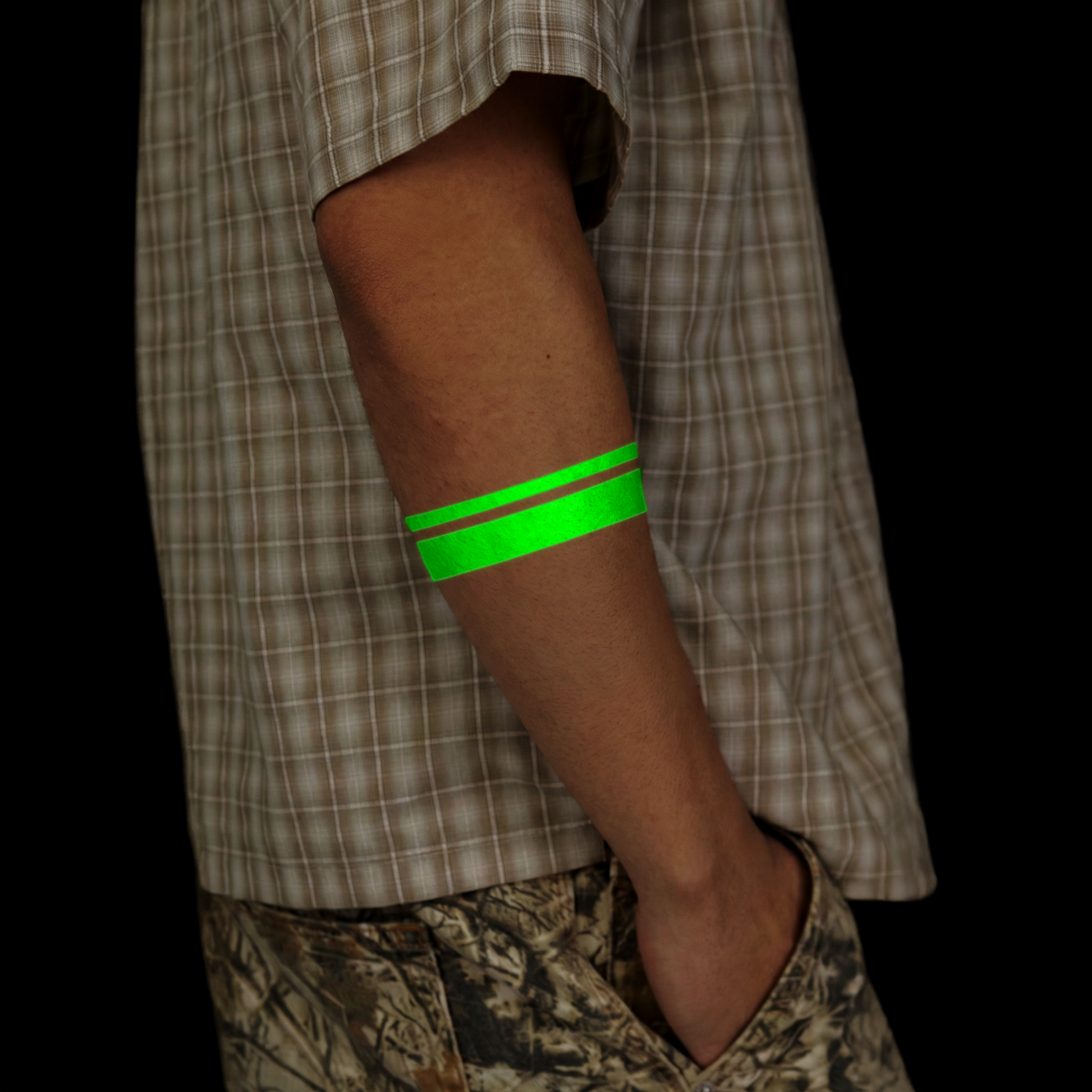 Person wearing a green arm band with a plaid shirt and camouflage pants on a black background