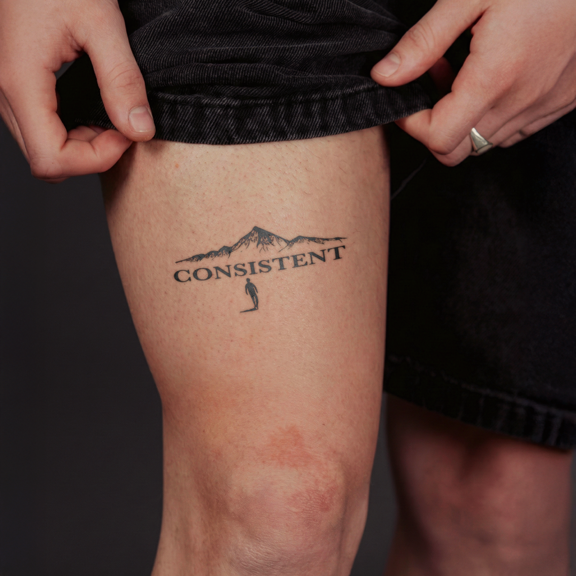 Tattoo of a mountain and the word 'CONSISTENT' on a person's leg.