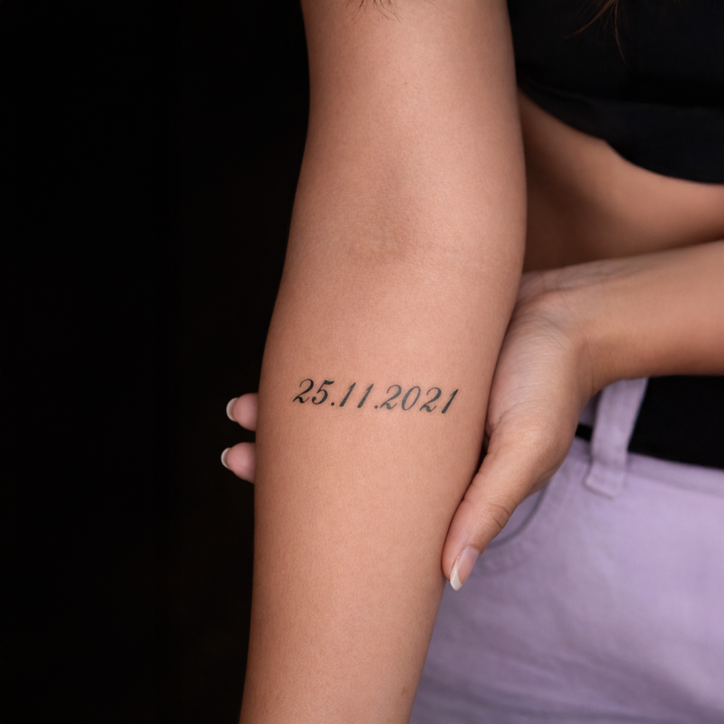 Person with a tattoo on their arm displaying '25.11.2021' against a black background