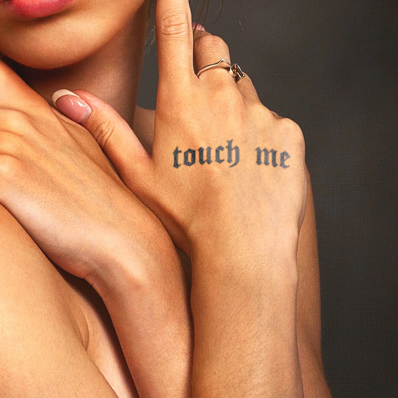 Close-up of a hand with 'touch me' tattoo on a dark background