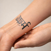 Tattoo of a stylized letter 'H' on a person's wrist with a neutral background