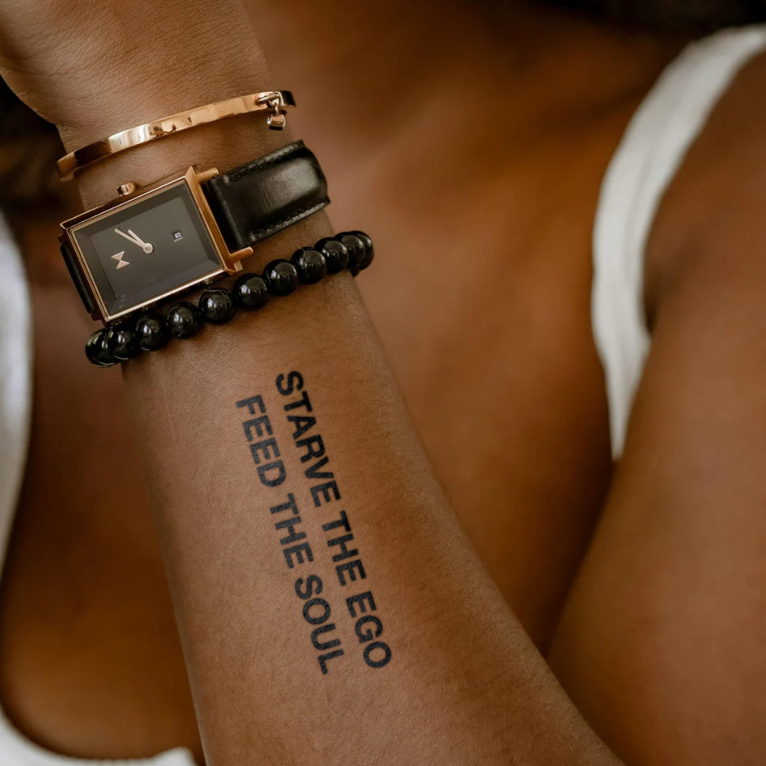 Person wearing a watch with a tattoo on their arm that reads 'Starve the Ego Feed the Soul'.