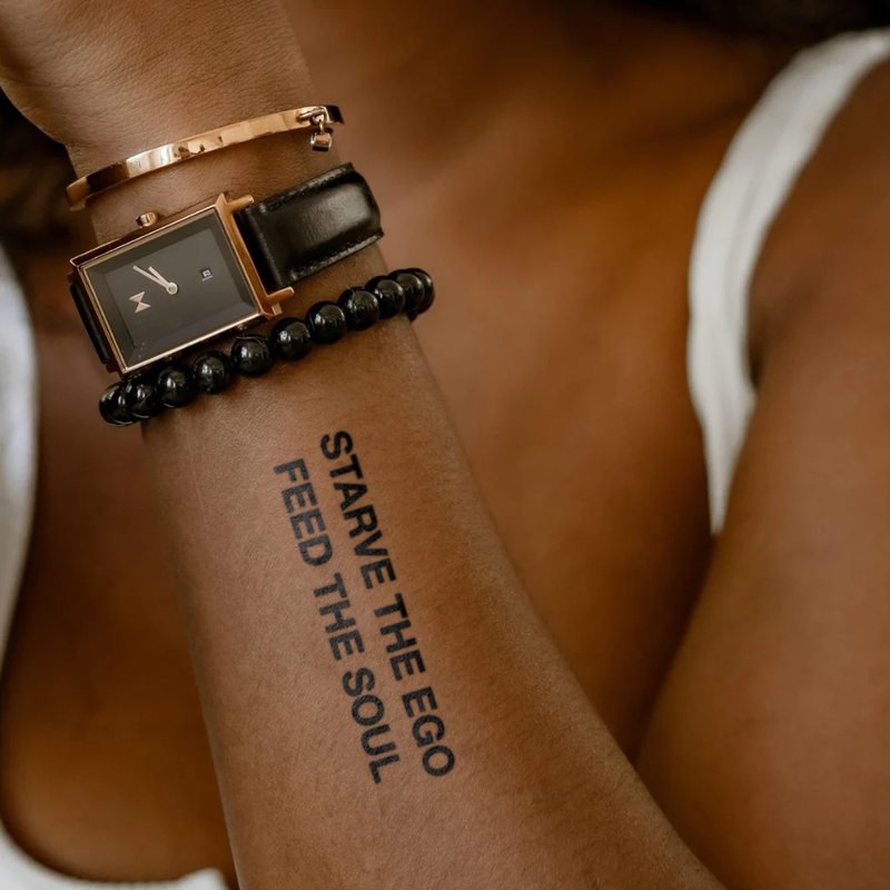 Person wearing a watch with a tattoo on their arm that reads 'Starve the Ego Feed the Soul'.