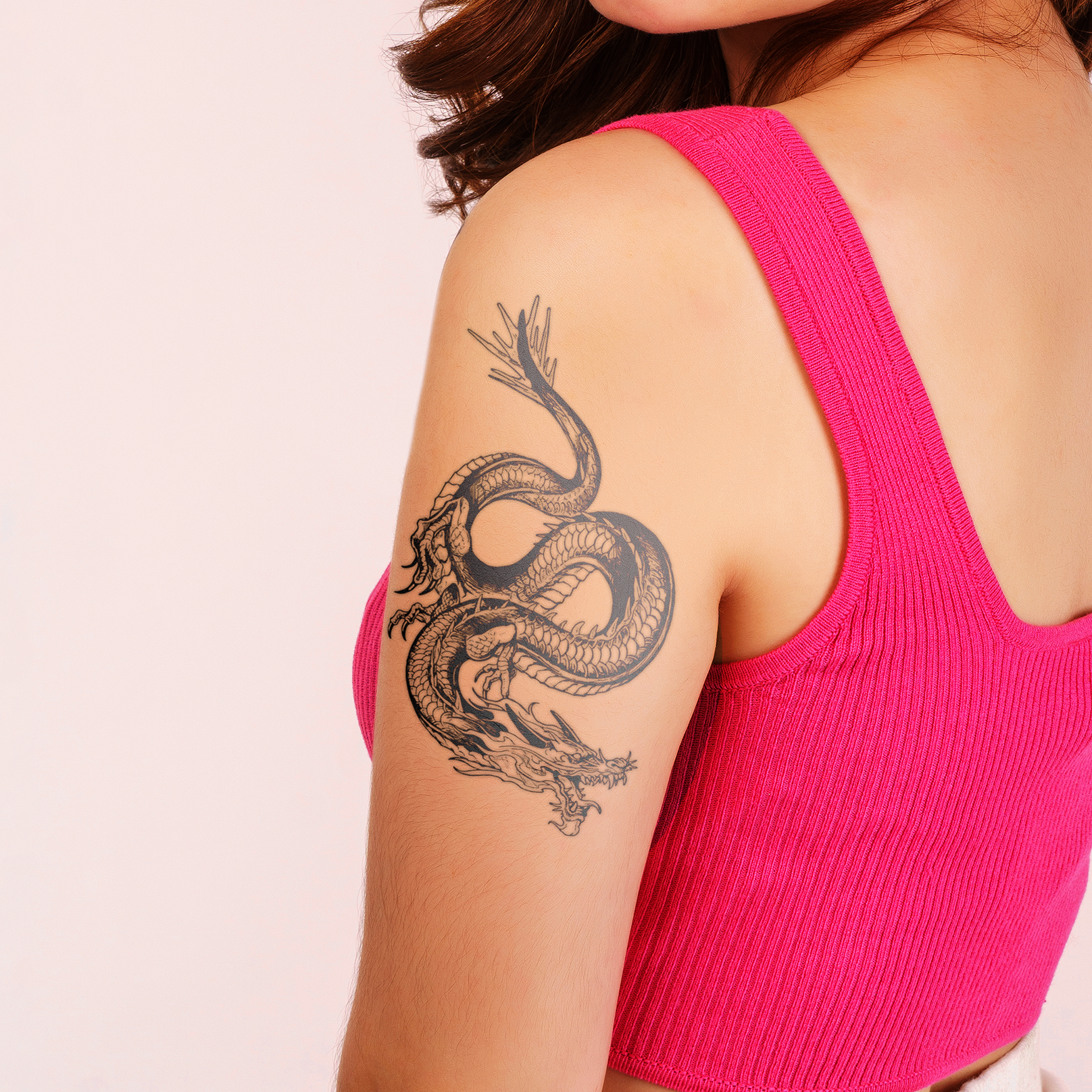 Person wearing a pink tank top with a dragon tattoo on their arm against a light pink background