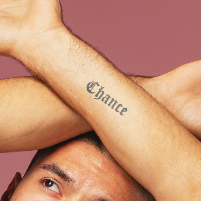 Person with 'Chance' tattoo on arm against a pink background