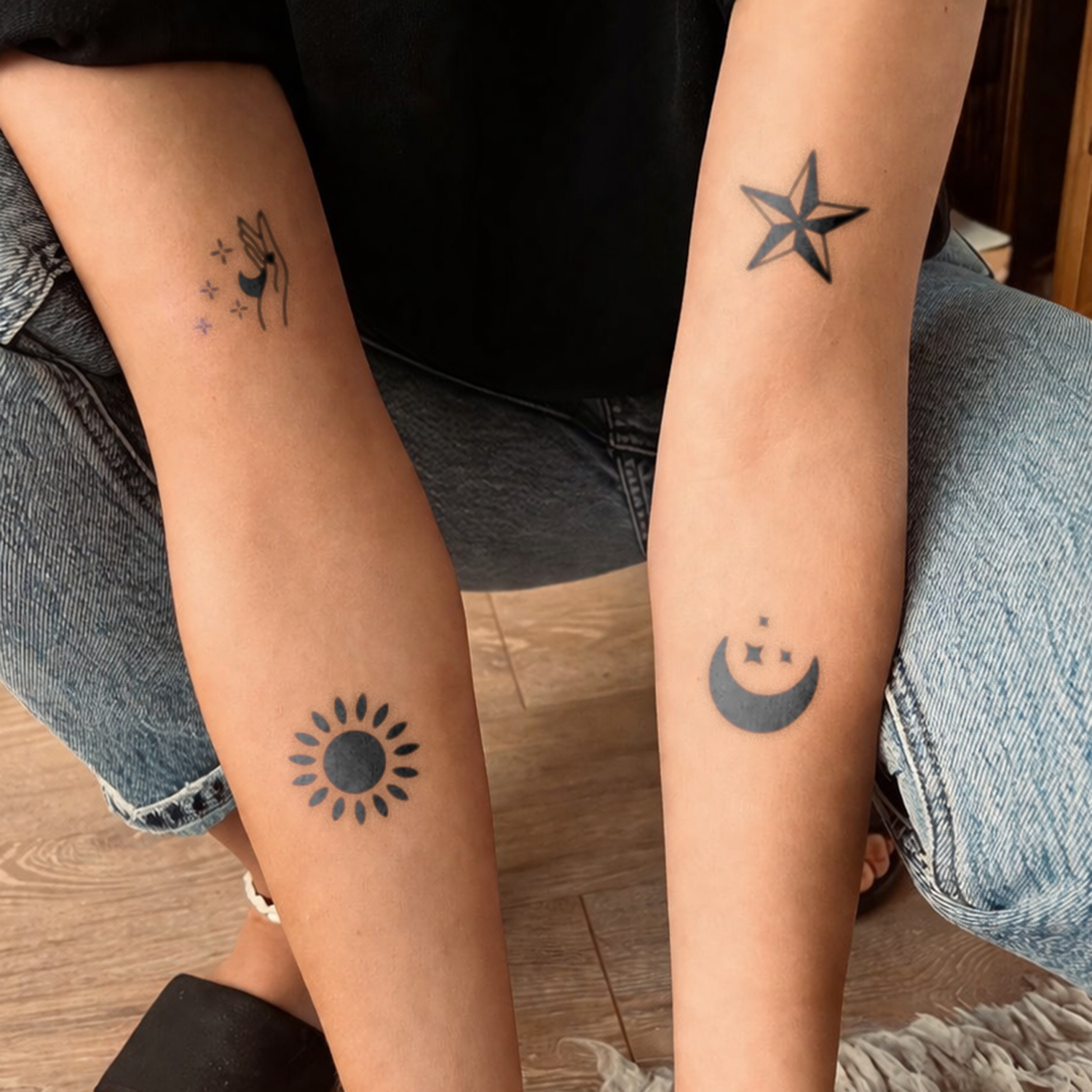 Two arms with tattoos of a star, sun, and moon on a wooden floor.