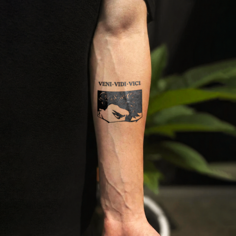 Tattoo on a person's arm with 'VENI VIDI VICI' and a face design, against a dark background.