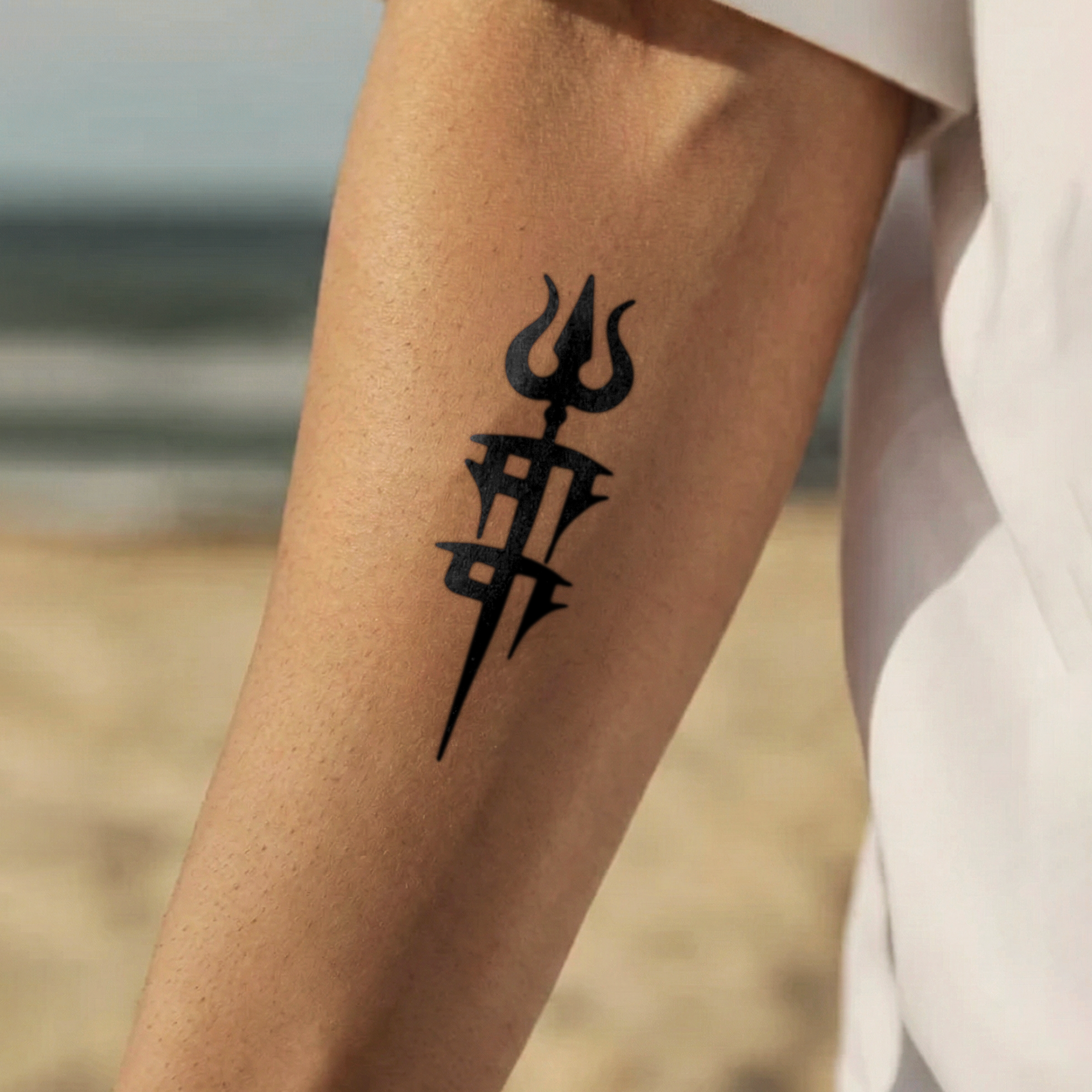 Black trident tattoo on a person's arm with a blurred beach background