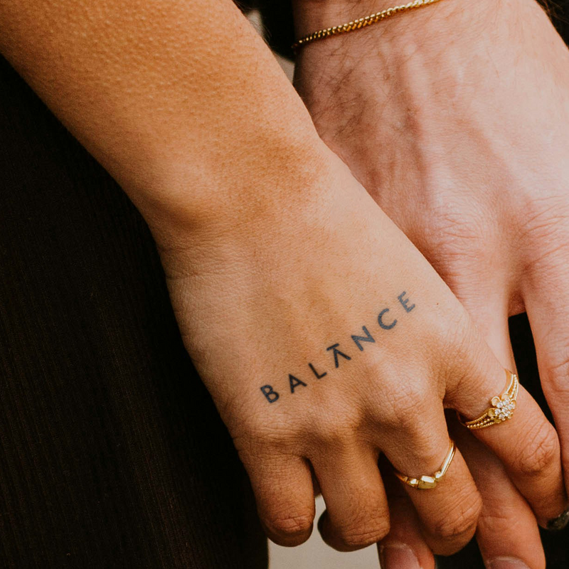 Hand with 'BALANCE' tattoo holding another hand, wearing gold rings.