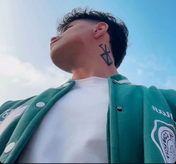 Person wearing a green jacket with a white shirt, standing against a blue sky.